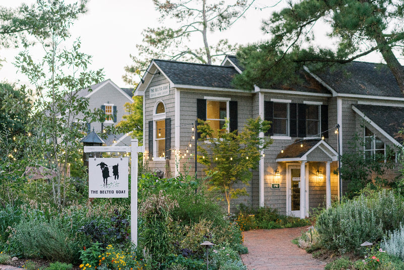 A charming gray house with white trim sits amid lush gardens and string lights. A sign in front, featuring two goats, reads “The Betting Goat.” A brick pathway leads to the entrance under tall trees.