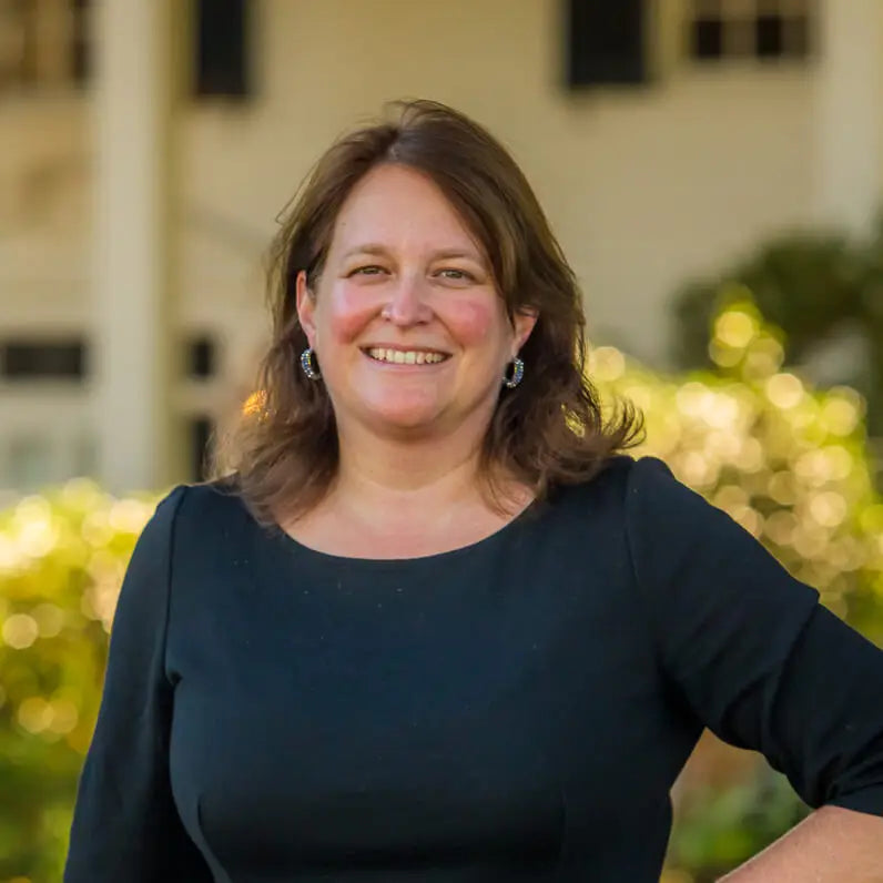 A woman with brown hair in a black dress smiles outdoors with greenery and a building in the background.
