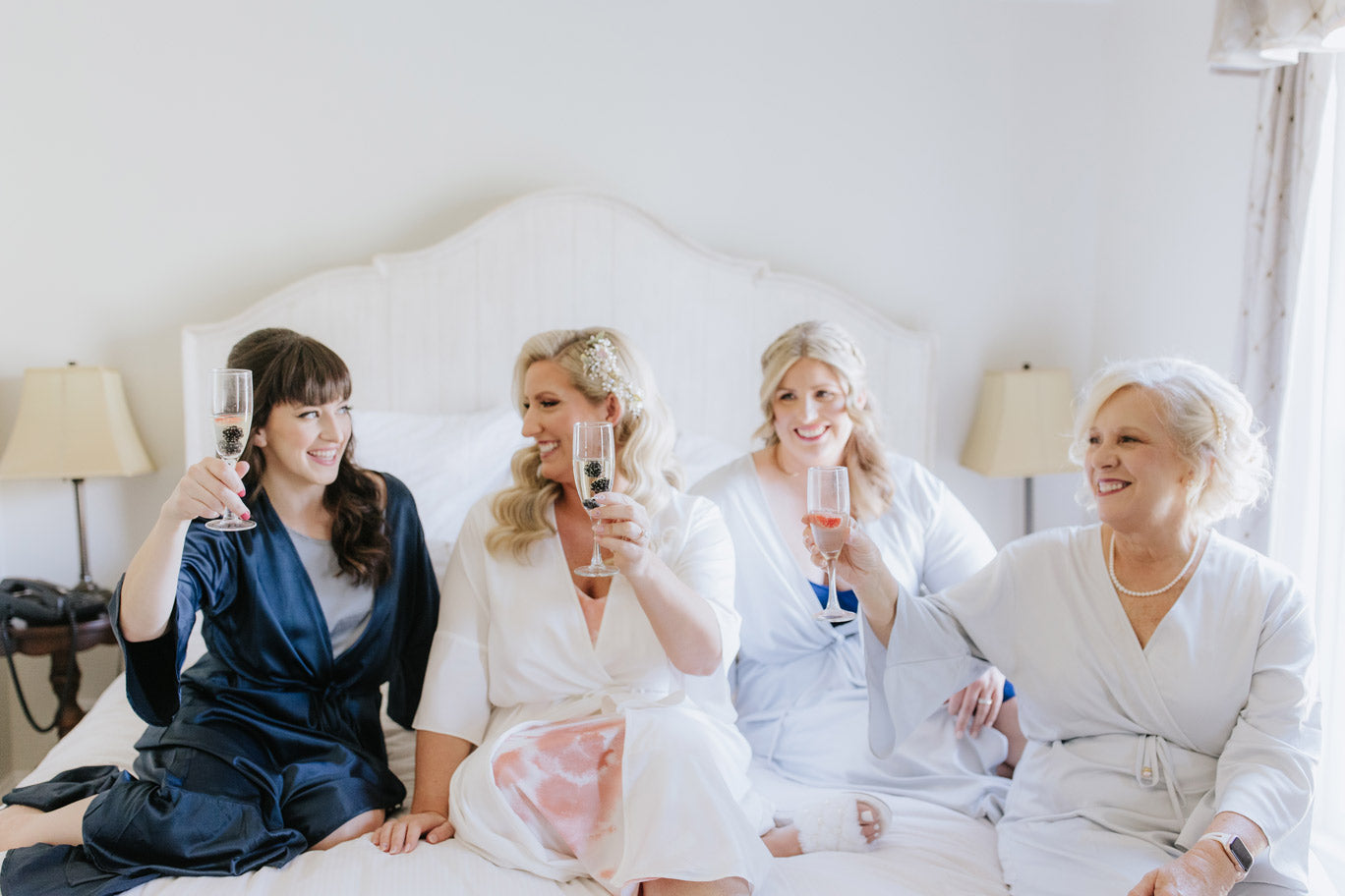 Four women in robes sit on a bed, smiling and raising glasses of pink champagne in a toast. The room is bright with soft natural light, and the mood appears joyful and celebratory.