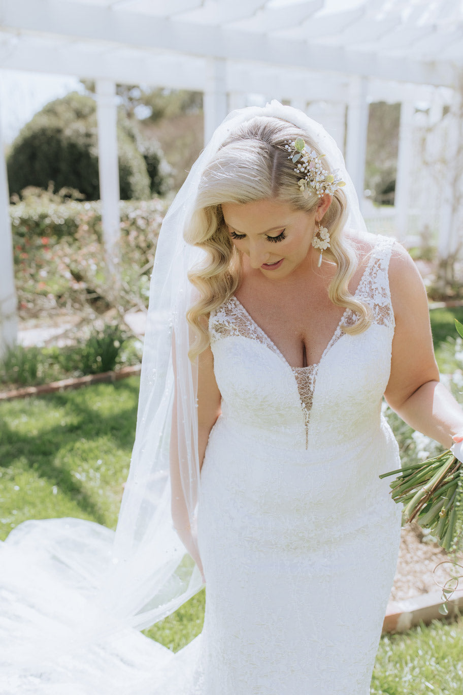 A bride in a white lace wedding dress and veil stands outdoors, holding part of her bouquet, with sunlight highlighting her curled blonde hair and floral hairpiece. Greenery and white columns are visible in the background.