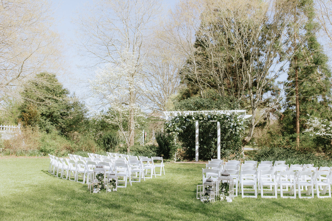 An outdoor wedding ceremony setup features two rows of white chairs facing a white arbor decorated with greenery and flowers, set on green grass with trees in the background under a clear sky.