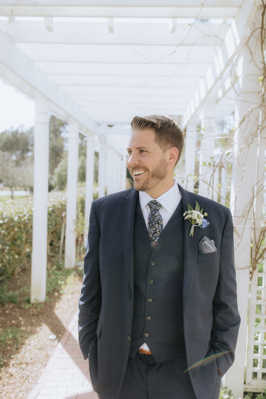 A man in a navy suit with a floral tie and boutonniere smiles while standing under a white pergola in a garden setting. Sunlight filters through the structure, creating a bright and airy atmosphere.
