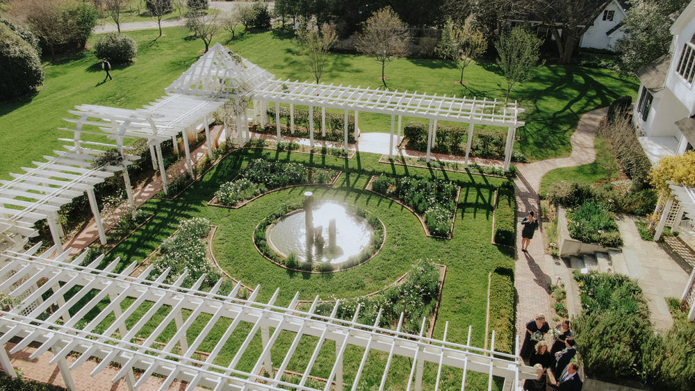 Aerial view of a formal garden with a circular pond at the center, surrounded by green lawns, white pergolas, pathways, and several people gathered near the garden entrance.