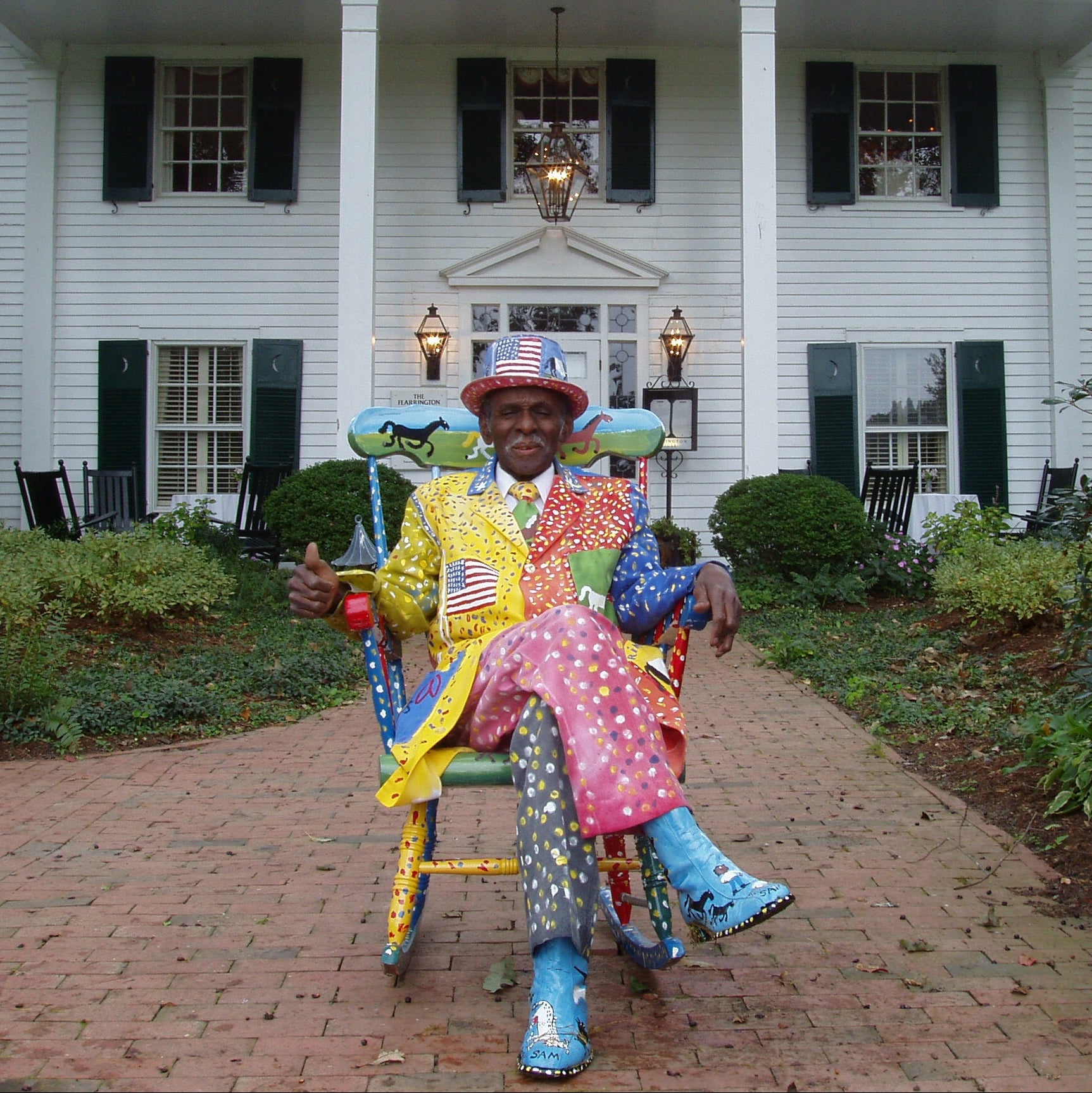 A man in a brightly colored, patterned suit and matching hat sits in a painted rocking chair on a brick path in front of a large white house with green shutters and a columned porch.