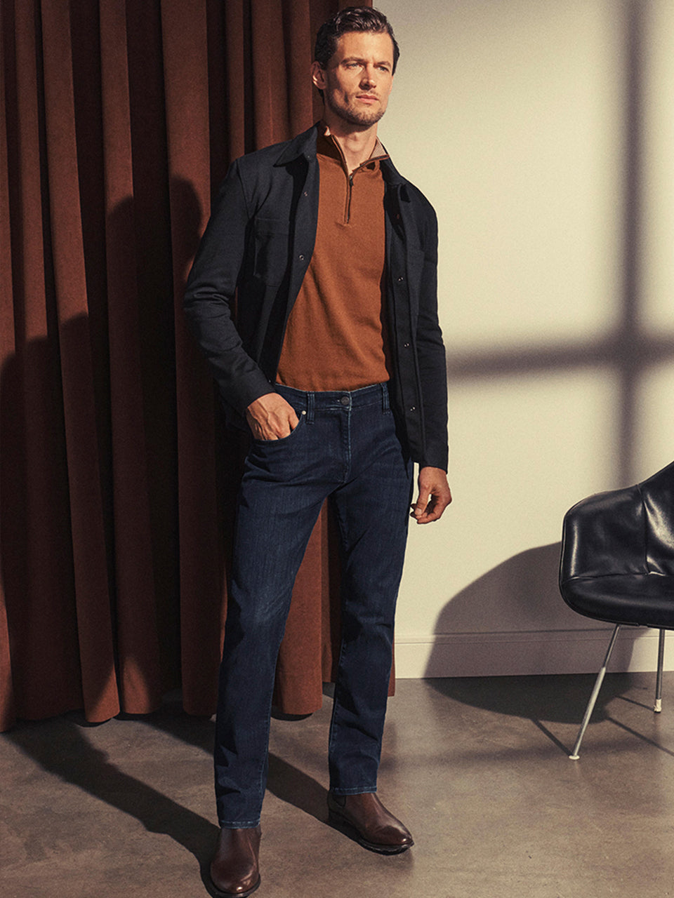 A man stands indoors near brown curtains and a black chair, wearing a black jacket over a brown polo shirt, dark blue jeans, and brown boots. Sunlight casts shadows on the wall and floor.