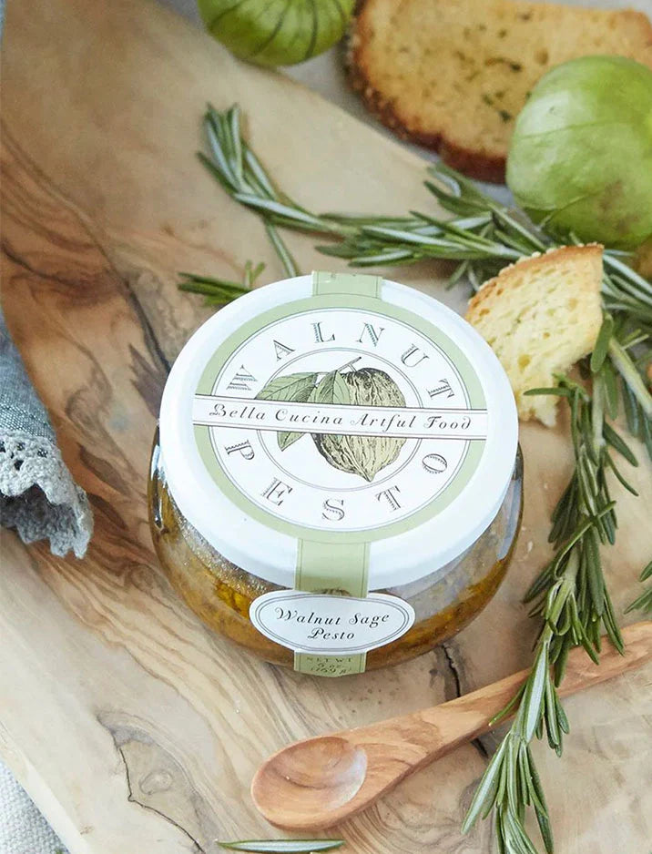 A jar of walnut sage pesto sits on a wooden board surrounded by sprigs of rosemary, slices of toasted bread, and a green tomatillo. The jar’s label reads “Walnut Pesto” and “Bella Cucina Artful Food.”.