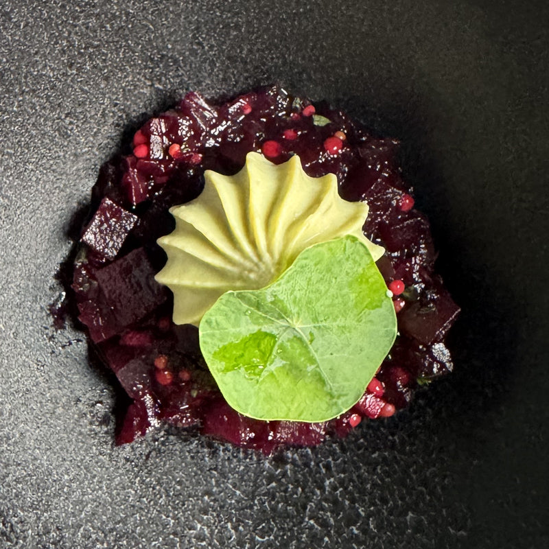 A gourmet dish featuring a mound of finely diced beets, topped with a swirl of creamy mousse and garnished with a single green leaf, served on a dark plate.