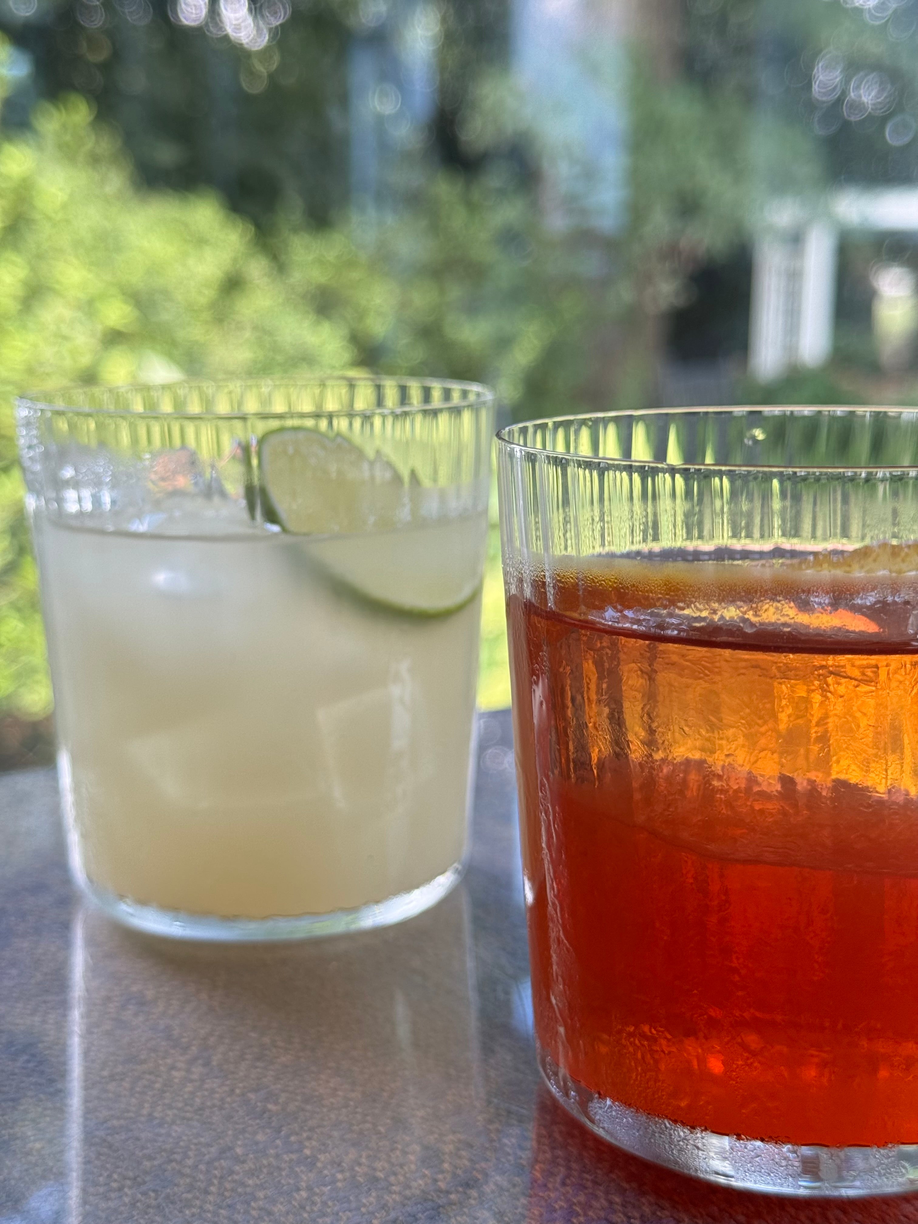 Two glasses on a table outdoors: one with a pale, lime-garnished drink and one with a bright orange beverage, both with ice, with a blurred green garden background.