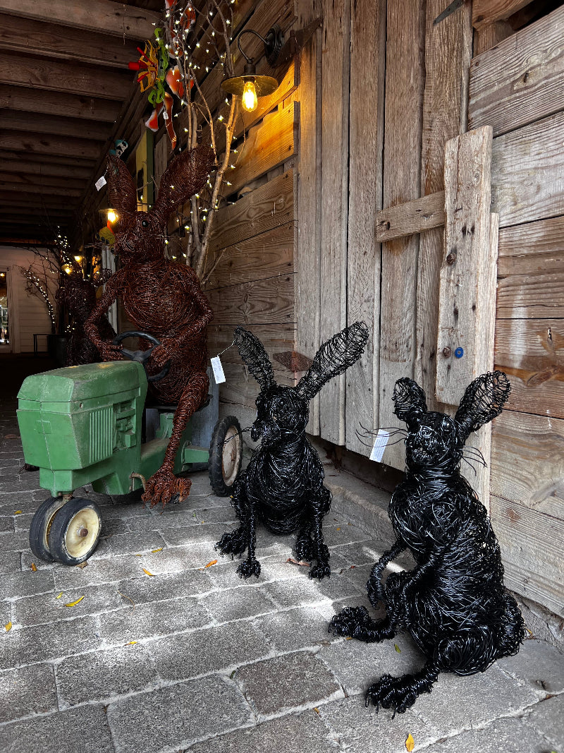 Two large black wire sculptures of rabbits sit on a stone path next to a rustic wooden wall, with another wire rabbit figure riding a green toy tractor in the background under string lights.