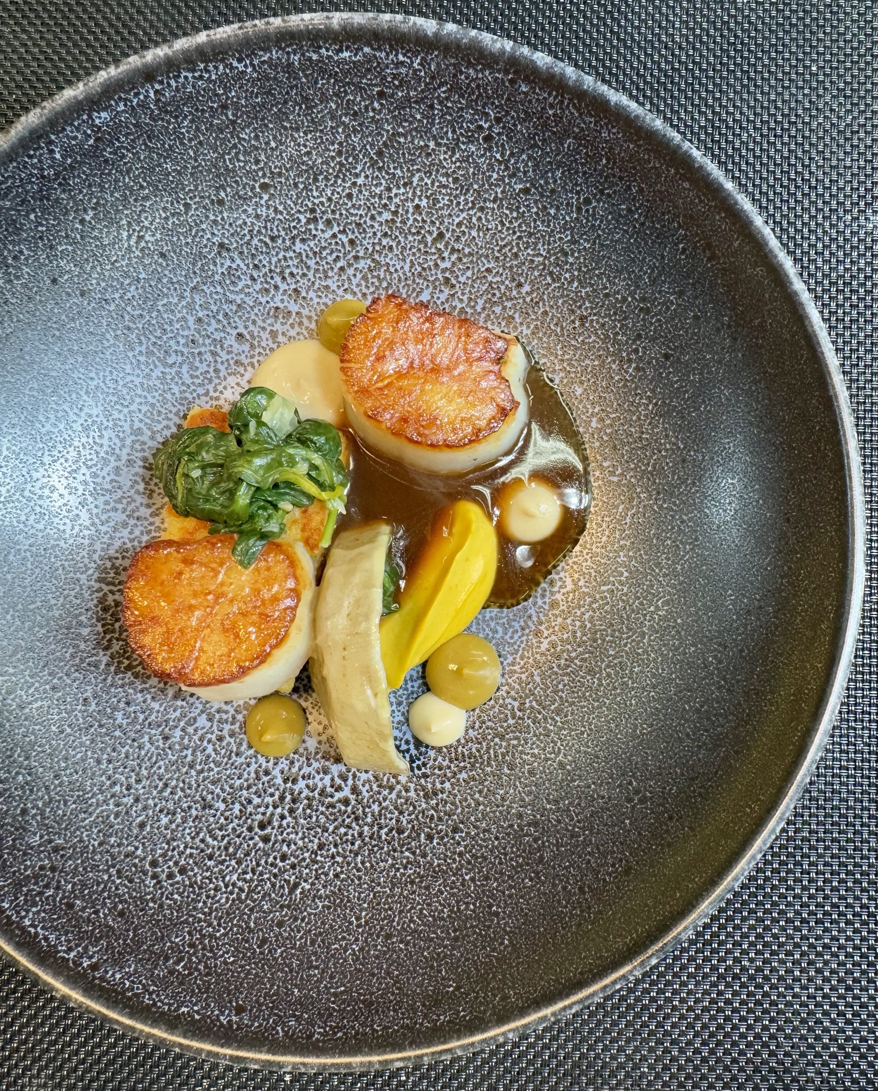 A gourmet dish featuring three seared scallops, colorful vegetable purées, leafy greens, and sauce, artistically arranged on a textured dark plate.