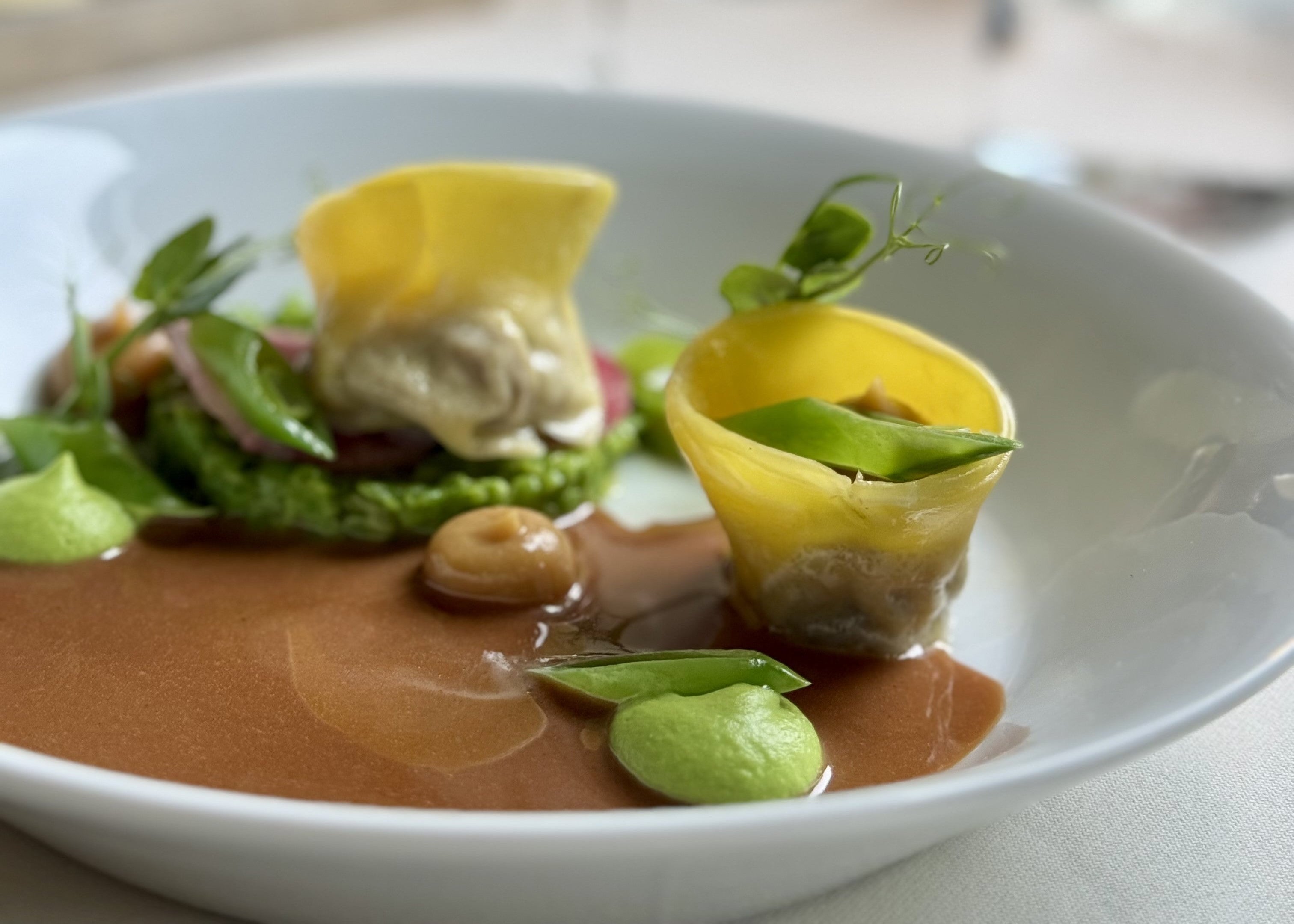 A white plate holds two gourmet stuffed pasta pieces with green puree, brown sauce, pea pods, and microgreens, artfully arranged for a fine dining presentation.