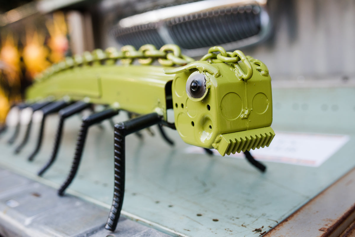 A green metal sculpture resembling a centipede, made from painted chain links and various mechanical parts, stands on a gray surface. The sculpture has black metal legs and a rectangular head with round features.