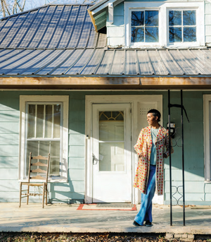 Person standing on a porch in front of a light blue house with a white door.