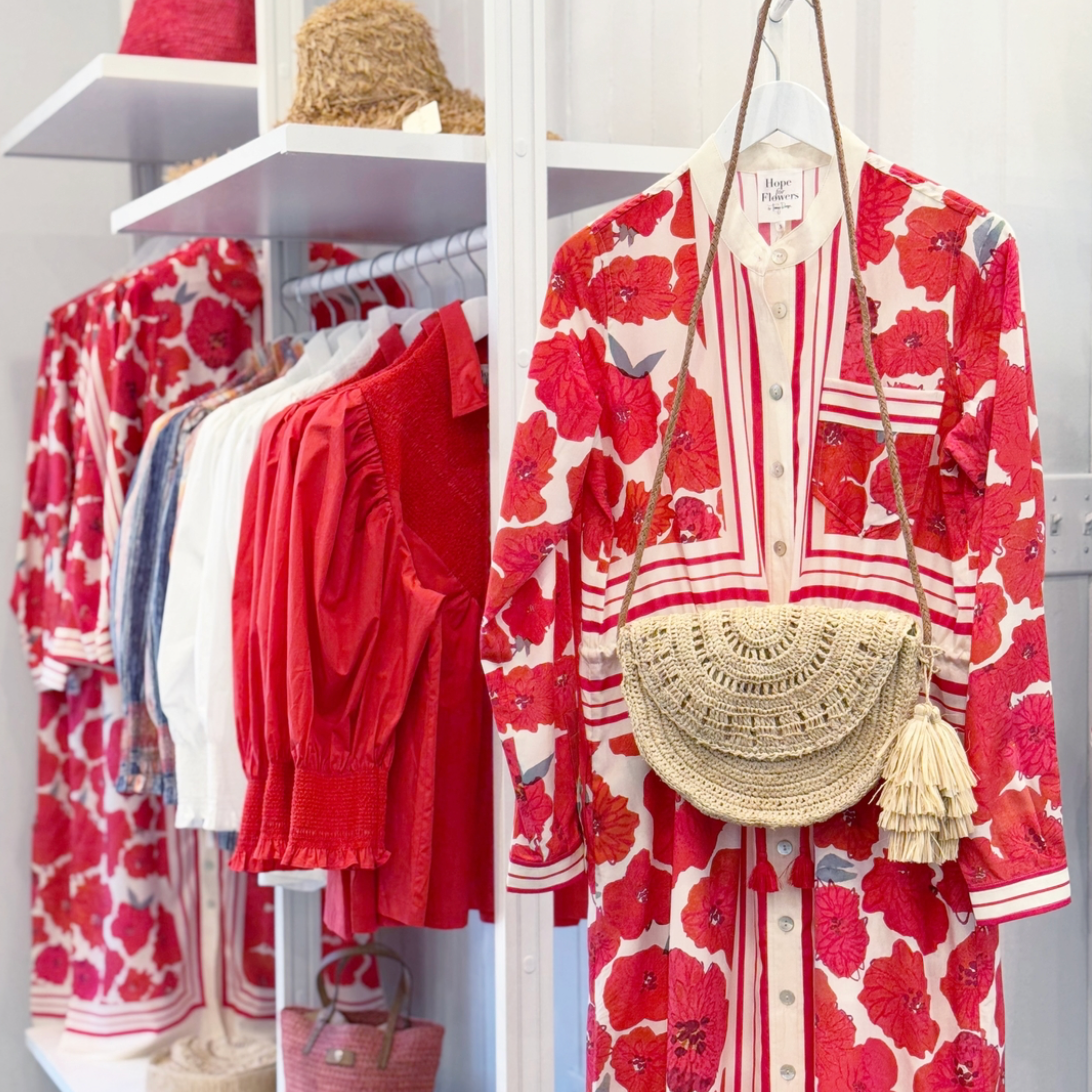 A clothing rack displays red and white floral dresses, a straw hat, and a woven straw handbag. A long, red floral dress with a straw bag hangs in the foreground, with more colorful garments behind it on white shelves.