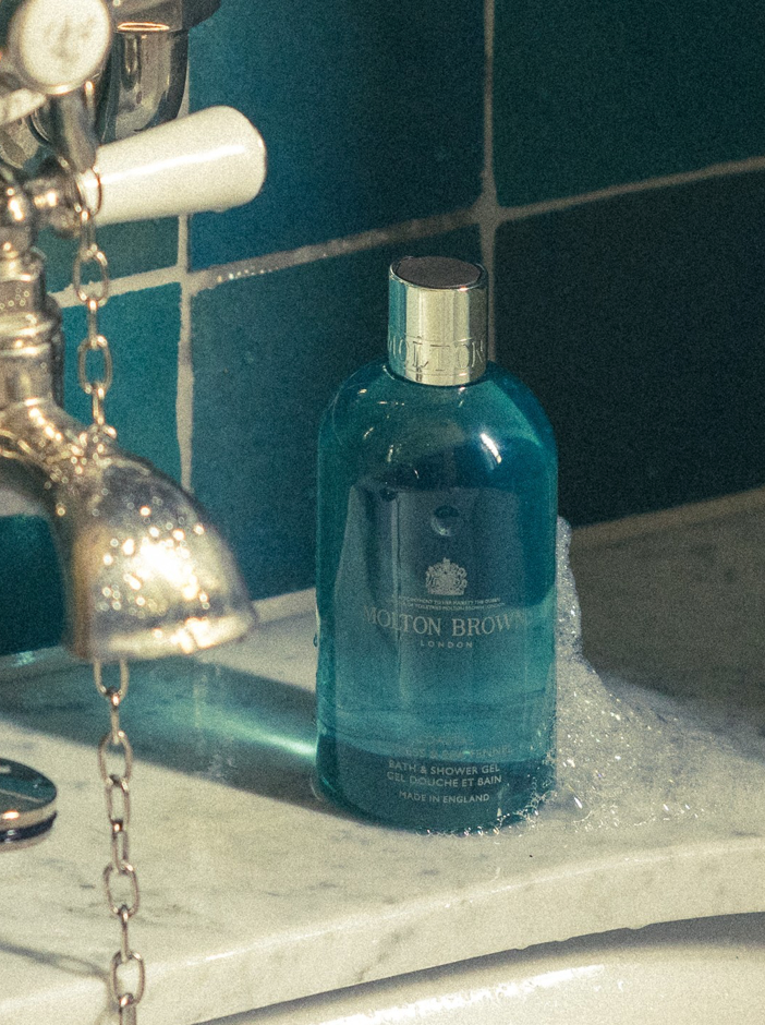 A bottle of Molton Brown London bath and shower gel sits on a marble edge next to a vintage bathtub faucet, partially covered in soap suds, with teal tiles in the background.