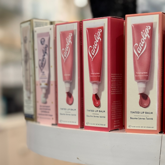 Close-up of several boxes of Lanolips Tinted Lip Balm displayed on a store shelf. The boxes are mostly pink and cream-colored with images of the lip balm tube on the front.