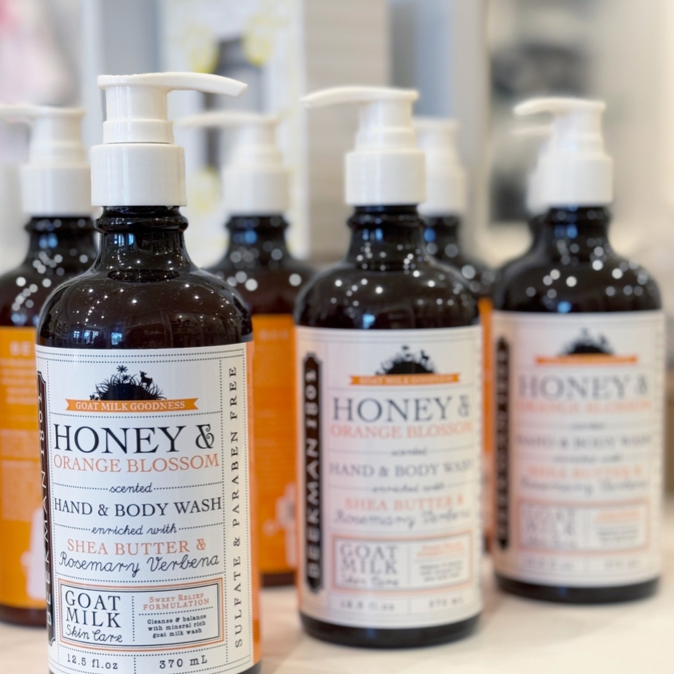 Several bottles of “Honey & Orange Blossom” hand and body wash with pump dispensers are displayed closely together. The labels mention goat milk, shea butter, rosemary, and that they are sulfate and paraben free.