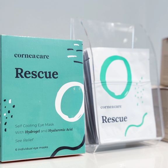 A box and individual packets of Cornea Care Rescue self cooling eye masks are displayed. The packaging is green and white with abstract designs and product details visible on the front.