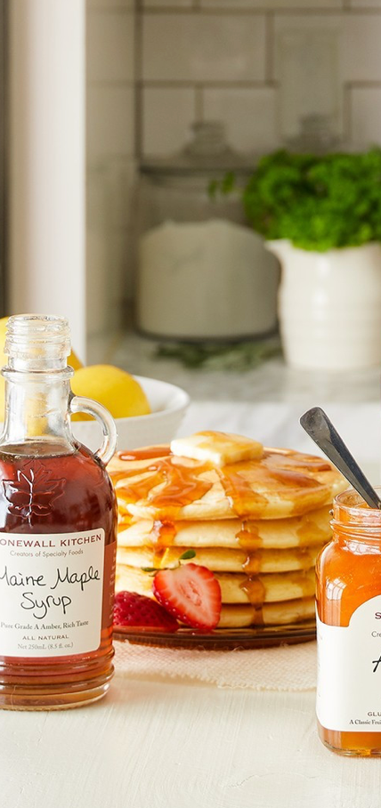 A stack of pancakes topped with syrup and sliced strawberries sits on a plate beside a bottle of maple syrup and a jar of orange marmalade on a light-colored kitchen table. A potted plant and lemons are in the background.