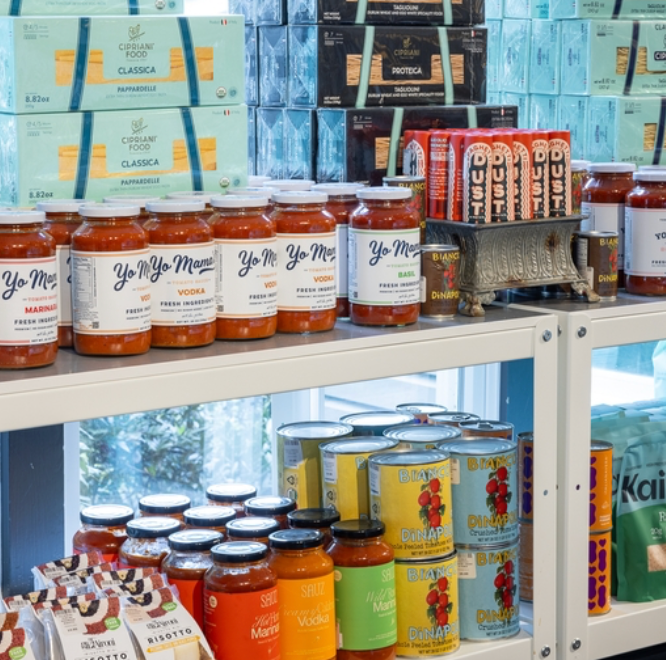 Shelves in a grocery store display jars of Yo Mama’s pasta sauce, boxes of pasta, cans of pineapple, and various packaged snacks arranged neatly with bright labels.