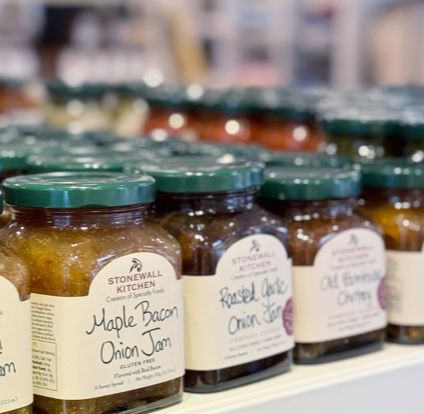 Jars of Stonewall Kitchen specialty jams, including Maple Bacon Onion Jam and Roasted Garlic Onion Jam, are neatly arranged on a store shelf with blurred jars in the background.