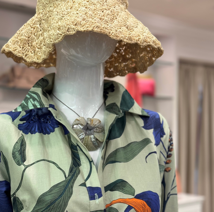 A mannequin dressed in a floral-patterned shirt and a large woven straw hat, accessorized with a flower-shaped pendant necklace, displayed in a brightly-lit clothing store.
