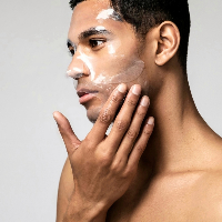 A shirtless man applies white cream to his face, using his hand to spread the product on his cheek. The background is plain and light-colored.