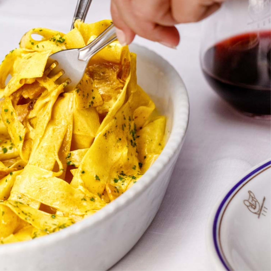 A hand uses a fork and spoon to twirl wide pasta noodles in a white dish, garnished with herbs. A glass of red wine and a white plate with a decorative rim are nearby on a white tablecloth.
