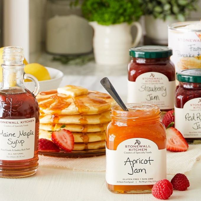 A breakfast table with a stack of pancakes topped with butter, surrounded by jars of Stonewall Kitchen Maine Maple Syrup, Apricot Jam, Strawberry Jam, Red Raspberry Jam, fresh strawberries, and pancake mix.