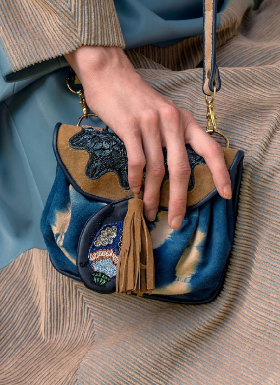 A close-up of a person's hand holding a small blue and beige handbag with embroidery, a brown tassel, and gold hardware. The person is wearing a textured blue and beige outfit.
