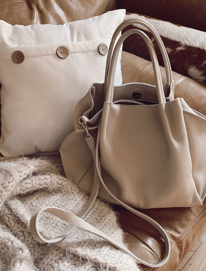 A beige leather handbag with a shoulder strap sits on a tan leather couch next to a buttoned white pillow and a chunky knit cream blanket.