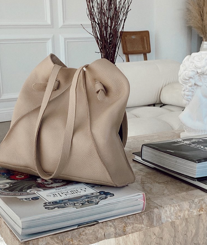 A beige leather tote bag sits on a marble coffee table next to stacked books in a modern living room with a white sofa, decorative vase, and neutral decor.