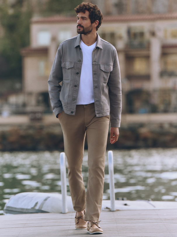 A man with dark hair and a beard walks outdoors by the water, wearing a light gray jacket over a white t-shirt, beige pants, and tan shoes. Blurred buildings and greenery are visible in the background.