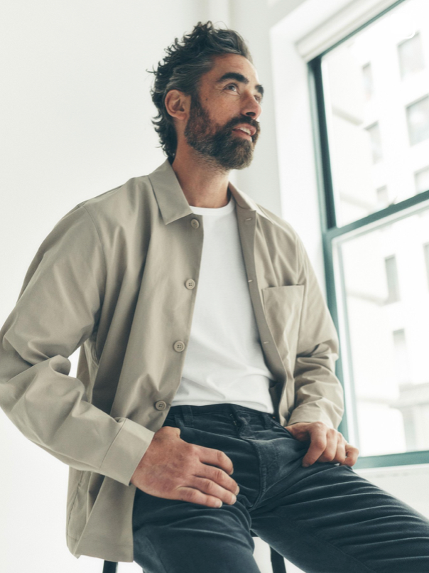 A man with dark hair and a beard sits on a stool indoors, wearing a light beige button-up shirt over a white T-shirt and black pants, looking to the side near a window with natural light.