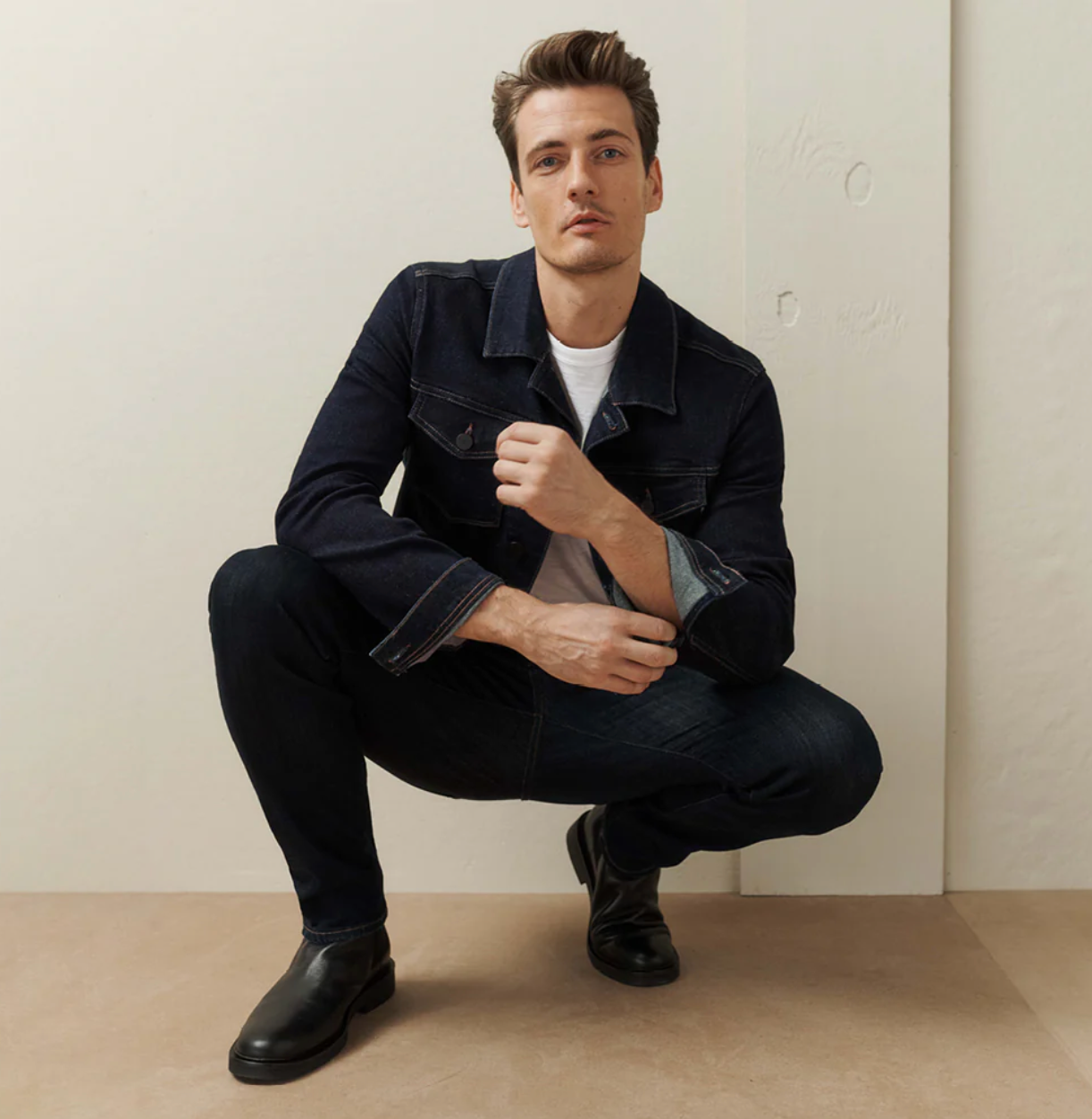 A man with short hair crouches indoors, wearing a dark denim jacket, dark jeans, black boots, and a white t-shirt, looking confidently at the camera against a minimal, neutral background.