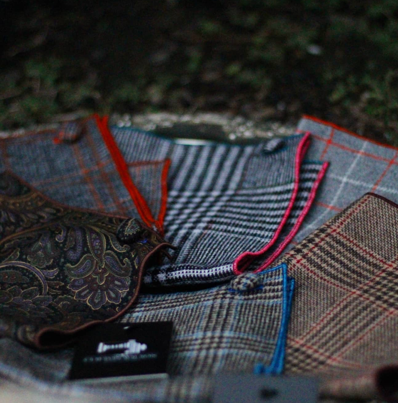 A close-up of several patterned and colorful tweed and paisley fabric swatches, neatly arranged outdoors on a surface, with blurred grass in the background.