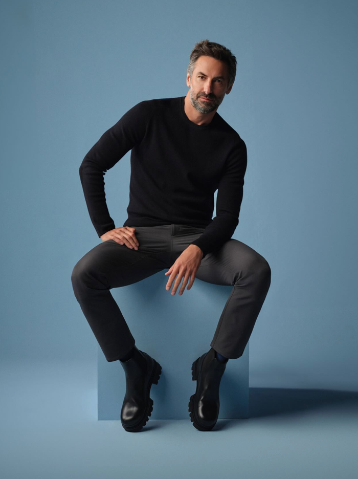 A man with short dark hair and a beard sits on a blue block against a blue background, wearing a black sweater, dark grey pants, and chunky black boots, looking confidently at the camera.