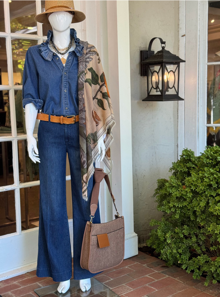 A mannequin dressed in a blue ruffled denim shirt, wide-leg jeans, tan belt, and hat. A floral scarf drapes over one shoulder, and a brown handbag hangs at the side. There is a lantern and green shrub nearby.