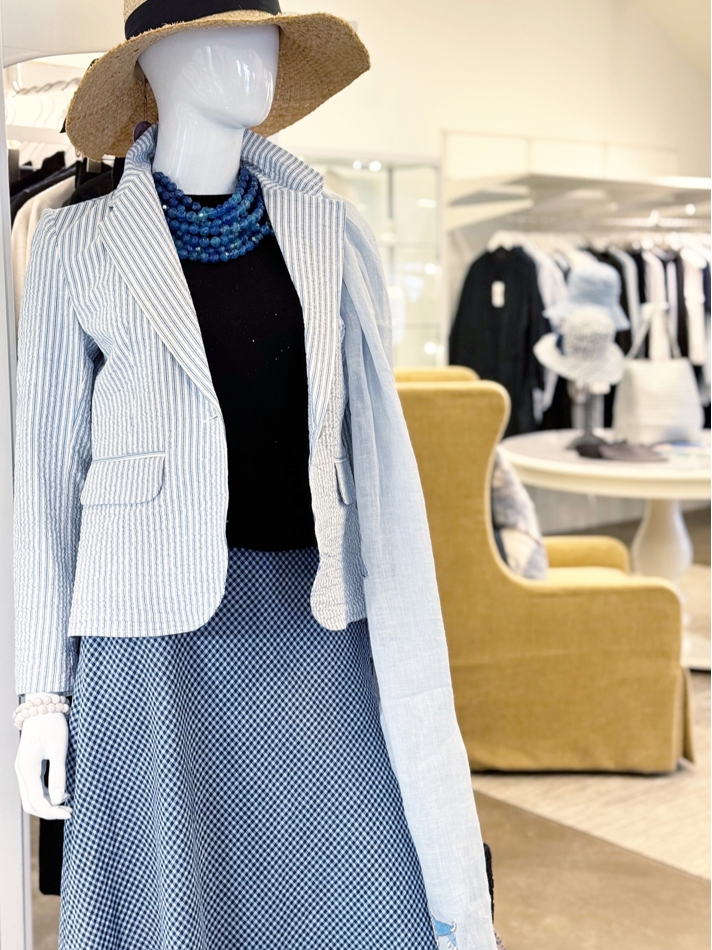 A mannequin dressed in a light blue pinstriped blazer, black top, blue patterned skirt, layered blue beads necklace, and a wide-brimmed straw hat in a bright, stylish boutique.