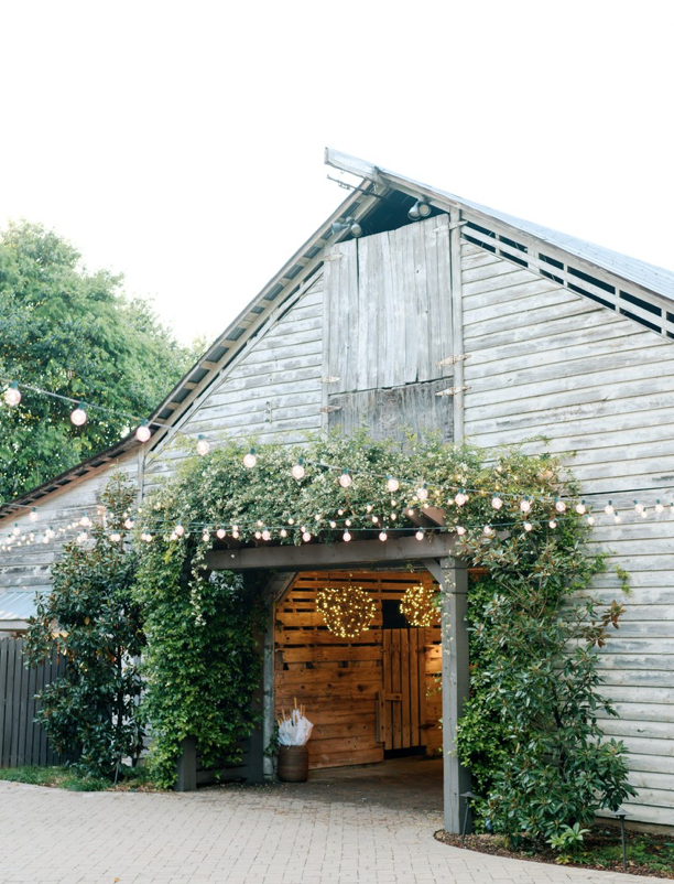 A rustic wooden barn decorated with string lights and greenery at the entrance. Warm lights inside and outside create a cozy, inviting atmosphere. The barn is surrounded by lush green trees and shrubs.
