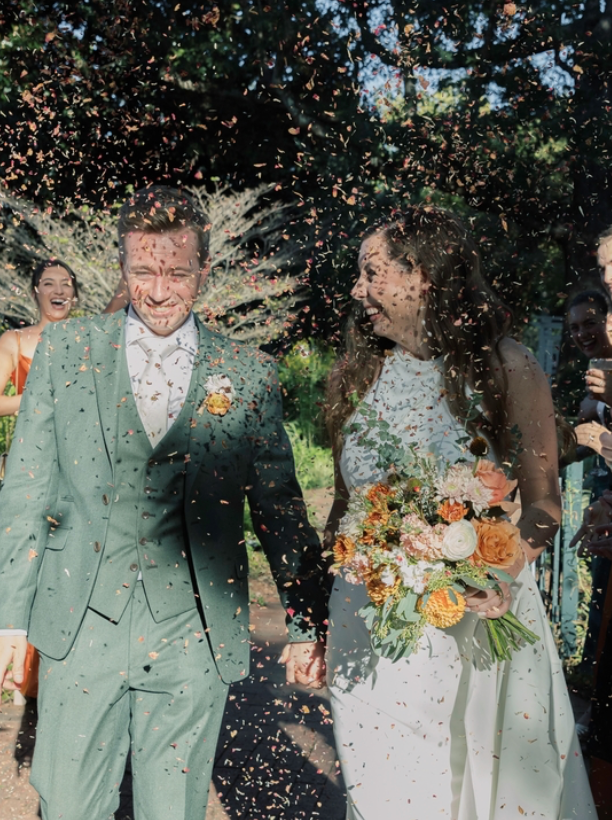 A newlywed couple, smiling and holding hands, walks outdoors as guests shower them with flower petals. The bride holds a colorful bouquet and wears a white dress; the groom is in a green suit. Sunlight filters through the trees.