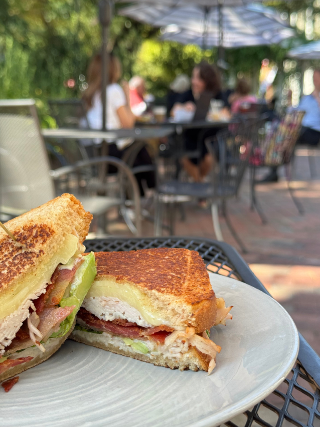 A close-up of a grilled turkey sandwich with cheese, bacon, and veggies on a white plate. In the blurred background, people sit at outdoor tables under umbrellas in a sunny garden patio.