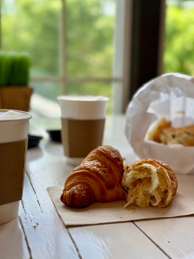 A close-up of a croissant, one half torn open, on a napkin with two takeaway coffee cups and a paper bag with more pastries in the background on a sunlit white table. Greenery is visible through the window behind.