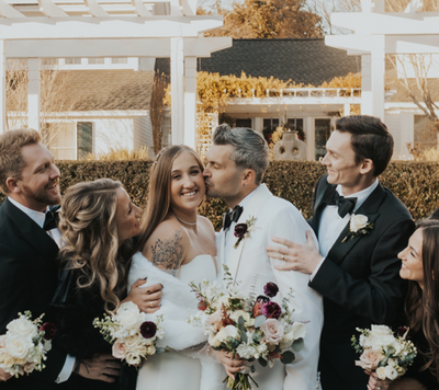 A bride and groom, both smiling, stand surrounded by their wedding party. The groom kisses the bride on the cheek as friends laugh, holding bouquets. They are dressed formally, with greenery and a white pergola behind them.
