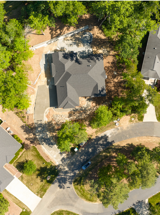 Aerial view of a house under construction surrounded by trees, with a curved driveway and nearby road. Neighboring homes and green spaces are also visible.