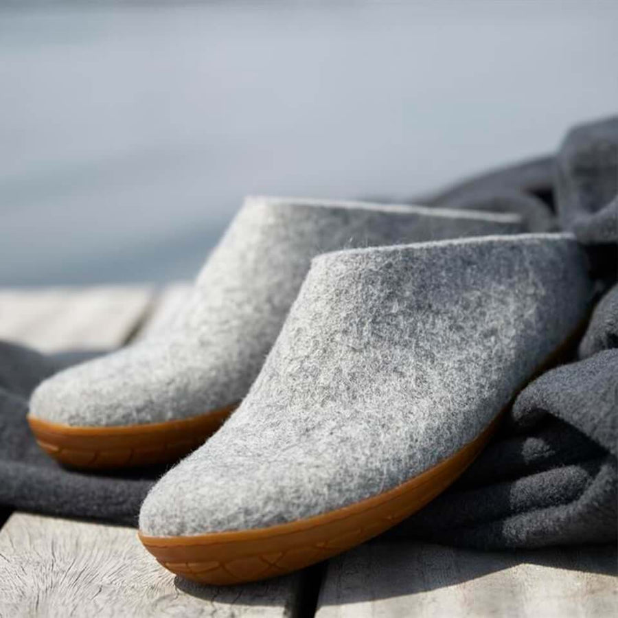 A pair of gray felt slippers with brown rubber soles rests on a wooden surface, with a soft gray cloth next to them and a blurred outdoor background.