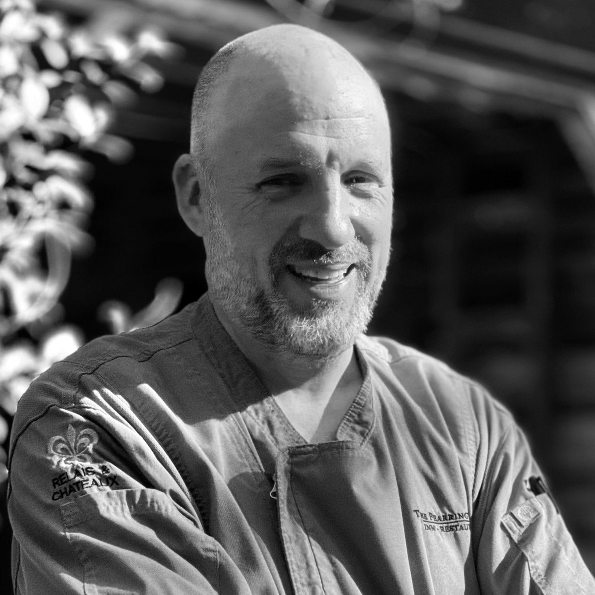 A bald man with a beard, wearing a chef’s jacket, stands outdoors with arms crossed, smiling. The image is in black and white, with blurred foliage and a building in the background.
