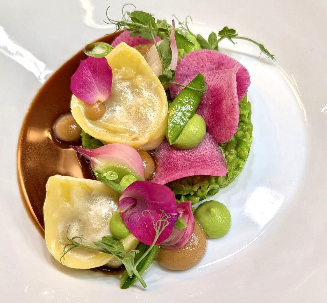 A gourmet dish featuring ravioli, green pea purée, pink radish slices, snap peas, edible flower petals, and a brown sauce artfully arranged on a white plate.