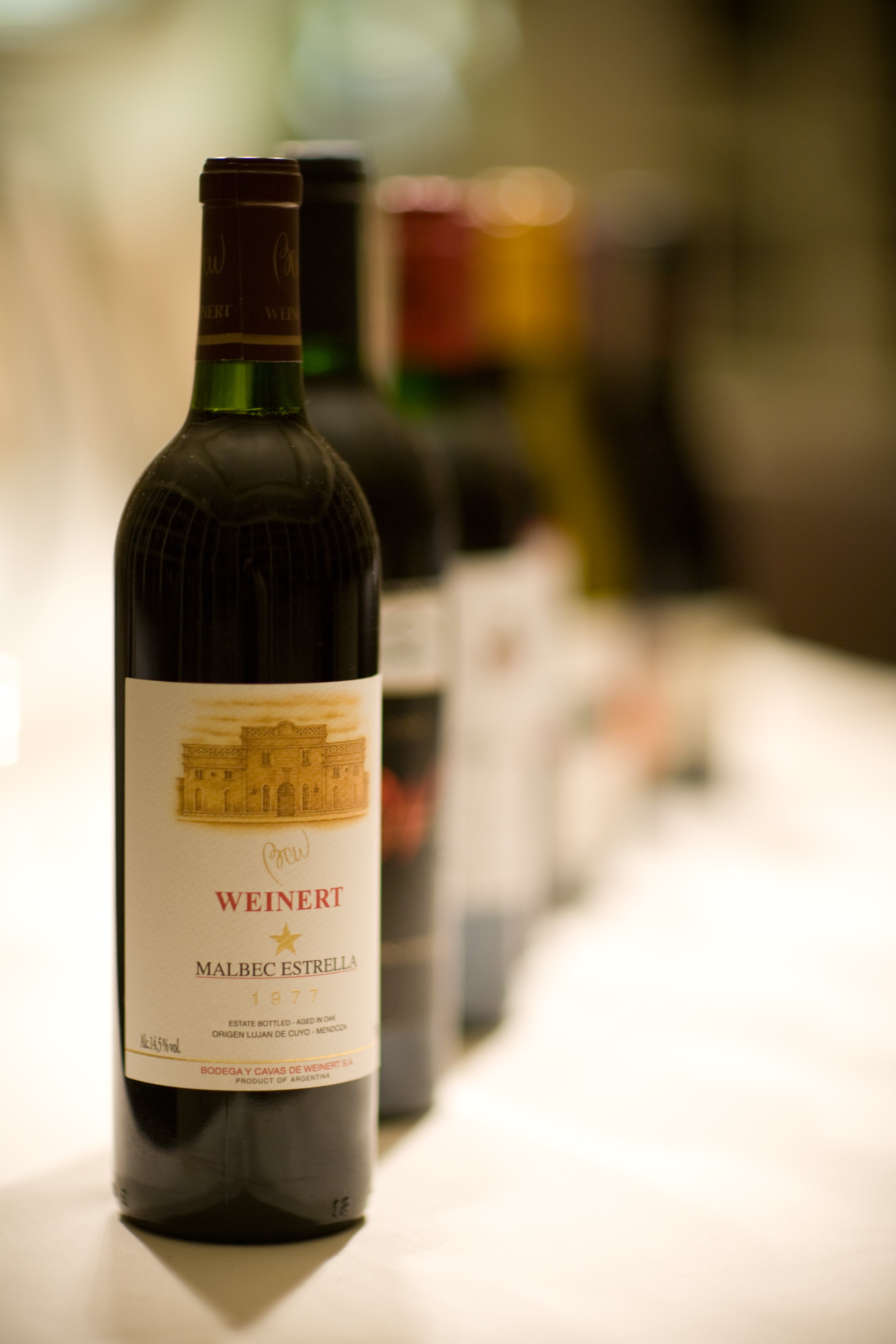 A bottle of Weinert Malbec Estrella red wine is in sharp focus at the front, with several other blurred wine bottles lined up behind it on a white surface.