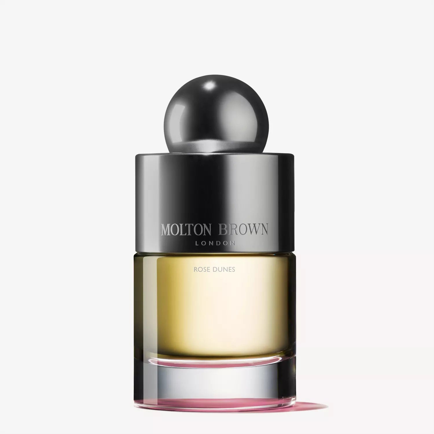 A clear glass bottle of MOLTON BROWN - ROSE DUNES EAU DE TOILETTE with a metallic cap and pink-tinted base, featuring velvety rose notes, is displayed against a plain white background.